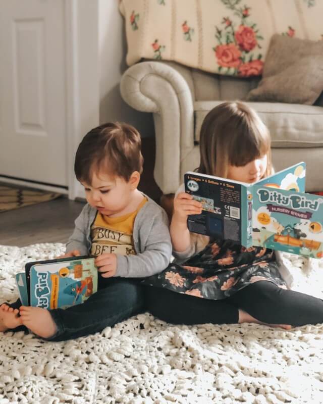 Two young children sit back to back on a light rug, each reading a book. Ditty Bird UK’s Nursery Rhymes educational toy rests nearby in the softly lit room, with a couch and floral blanket visible in the background.