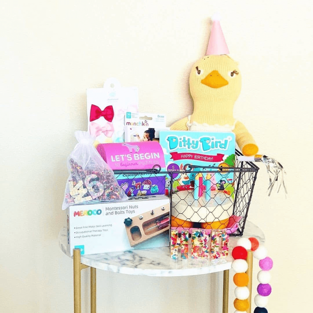 A table holds Ditty Bird UK's "Happy Birthday" basket with toys, books, toddler musical toys, candy, a plush duck in a pink party hat, beaded garland, and toy cake—the perfect first birthday gift before a plain white wall.