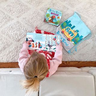 A child with blonde hair sits on a light carpet, enjoying Ditty Bird UK's Classical Ballet Music book—perfect for early learning. Nearby are another book and a blue drawstring bag featuring a city bus illustration.