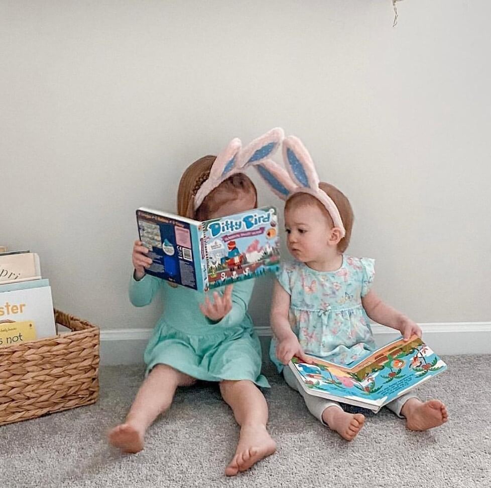 Two young children sit on a carpet, one in bunny ears holding Ditty Bird UK’s Classical Ballet Music book, while the other explores. A basket of books and an educational toy beside them makes it a perfect first birthday gift scene.