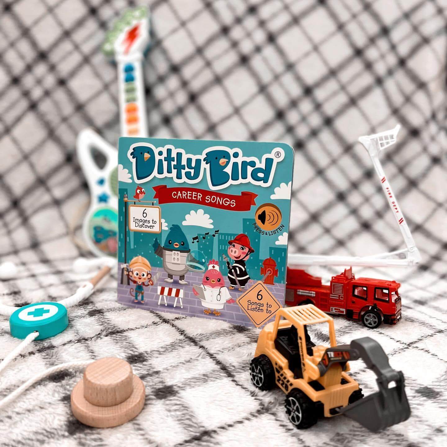 Ditty Bird UK’s Career Songs book is shown with toddler musical toys like a toy stethoscope, guitar, and wooden pieces on patterned fabric—an ideal educational toy and first birthday gift.