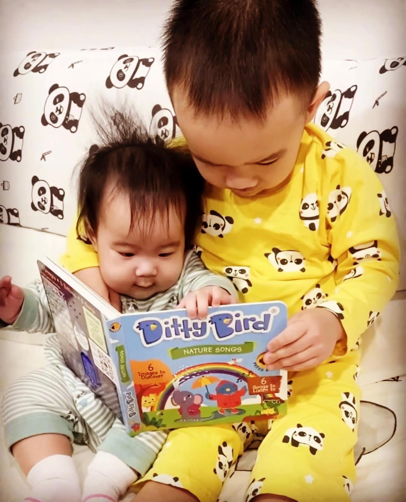 A young child in yellow panda pajamas reads "Nature Songs" by Ditty Bird UK to a baby in striped pajamas on a panda-patterned couch. Both children look engaged, making it an ideal early learning moment.