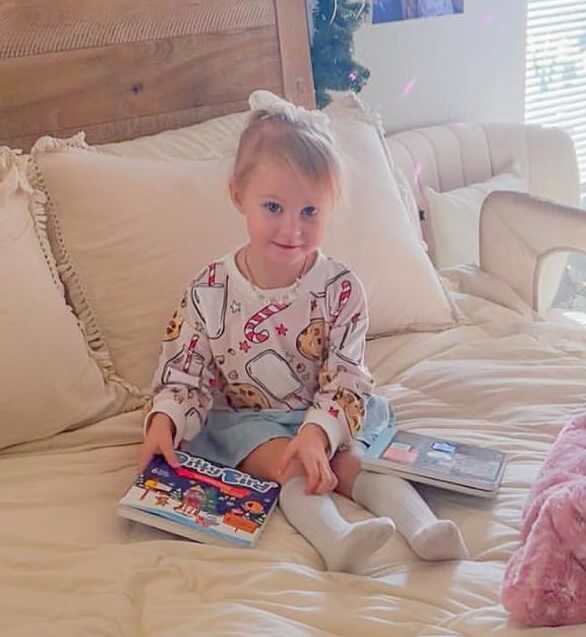 A young girl in a white bow sits on a bed, holding Ditty Bird UK's "Villancicos en Español" book—ideal for early learning. She wears a patterned white sweater, blue skirt, and knee-high socks, with another book beside her.