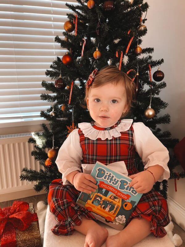 A young child in a red plaid dress sits by a decorated Christmas tree, holding Ditty Bird UK’s "Children's Songs" sound book. Wrapped presents and baby toys for ages 6–12 months are scattered on the floor nearby.