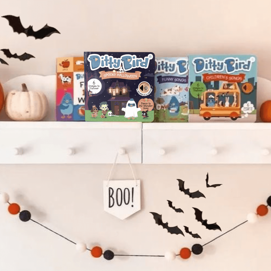 A white shelf with pumpkins, black paper bats, and three Ditty Bird UK “NEW! Spooky Halloween” interactive books. Below hangs a felt ball garland and a BOO! sign for a festive Halloween look featuring an educational toy for little ones.