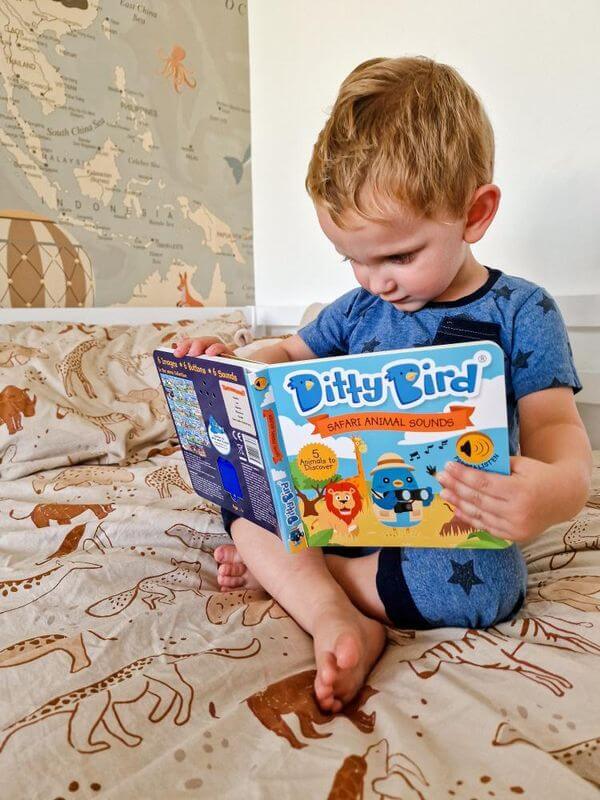 A young child sits cross-legged on animal-print sheets, absorbed in the Ditty Bird UK Safari Animal Sounds interactive book. With a world map on the wall, it makes an ideal early learning or first birthday gift.