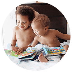 Two young children sit together on a bed, smiling as they explore colorful sound books. The older child holds a book open while the younger one looks on, both appearing happy and engaged by these early learning musical books.