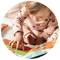 A young child in pink pajamas with black stars sits on a bed, intently looking at and touching the pages of a colorful sound book—one of the perfect gifts for one year old toddlers.
