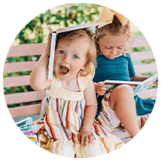 Two young children sit on a pink bench outdoors. One playfully balances a book on their head and smiles, while the other looks down at a story—perfect inspiration for early learning musical books or gifts for one year old.