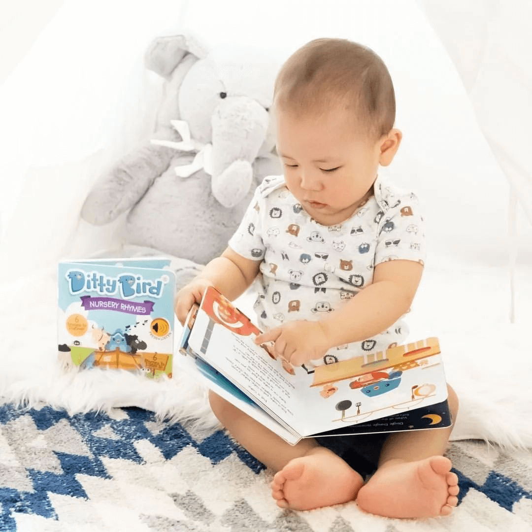 A baby sits on a patterned rug inside a white tent, looking at a book. Nearby is Ditty Bird UK's “Nursery Rhymes” educational toy—an ideal first birthday gift—and a plush elephant. The baby wears an animal print white shirt.