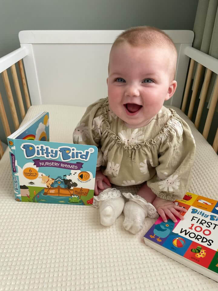 A smiling baby in a light green dress sits in a crib with Ditty Bird UK’s Nursery Rhymes interactive sound book—an educational toy perfect for babies 6-12 months.