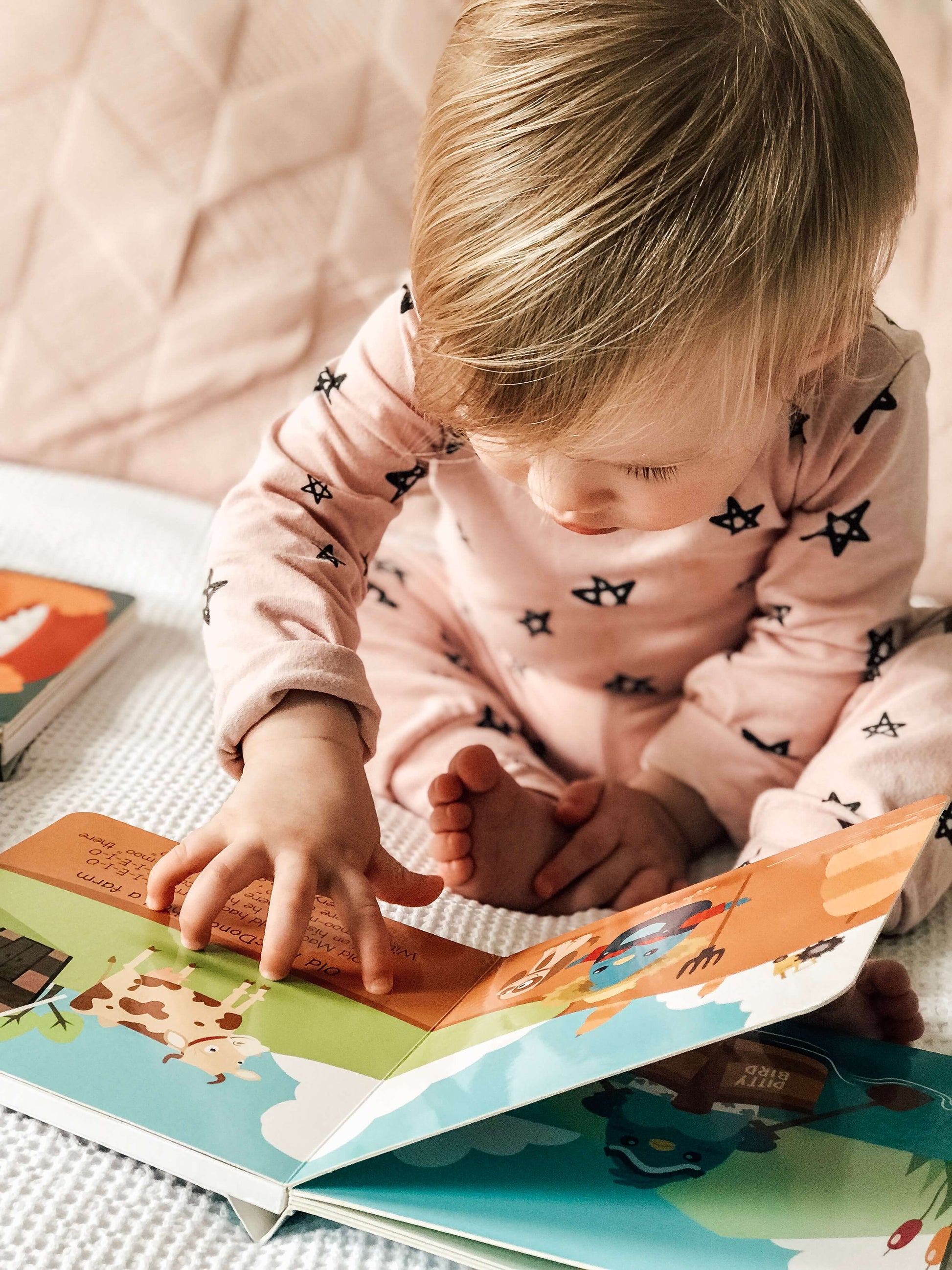 A young child in pink star pajamas sits on a bed, exploring the colorful pages of Ditty Bird UK's "Nursery Rhymes" book, surrounded by educational toys designed for early learning.