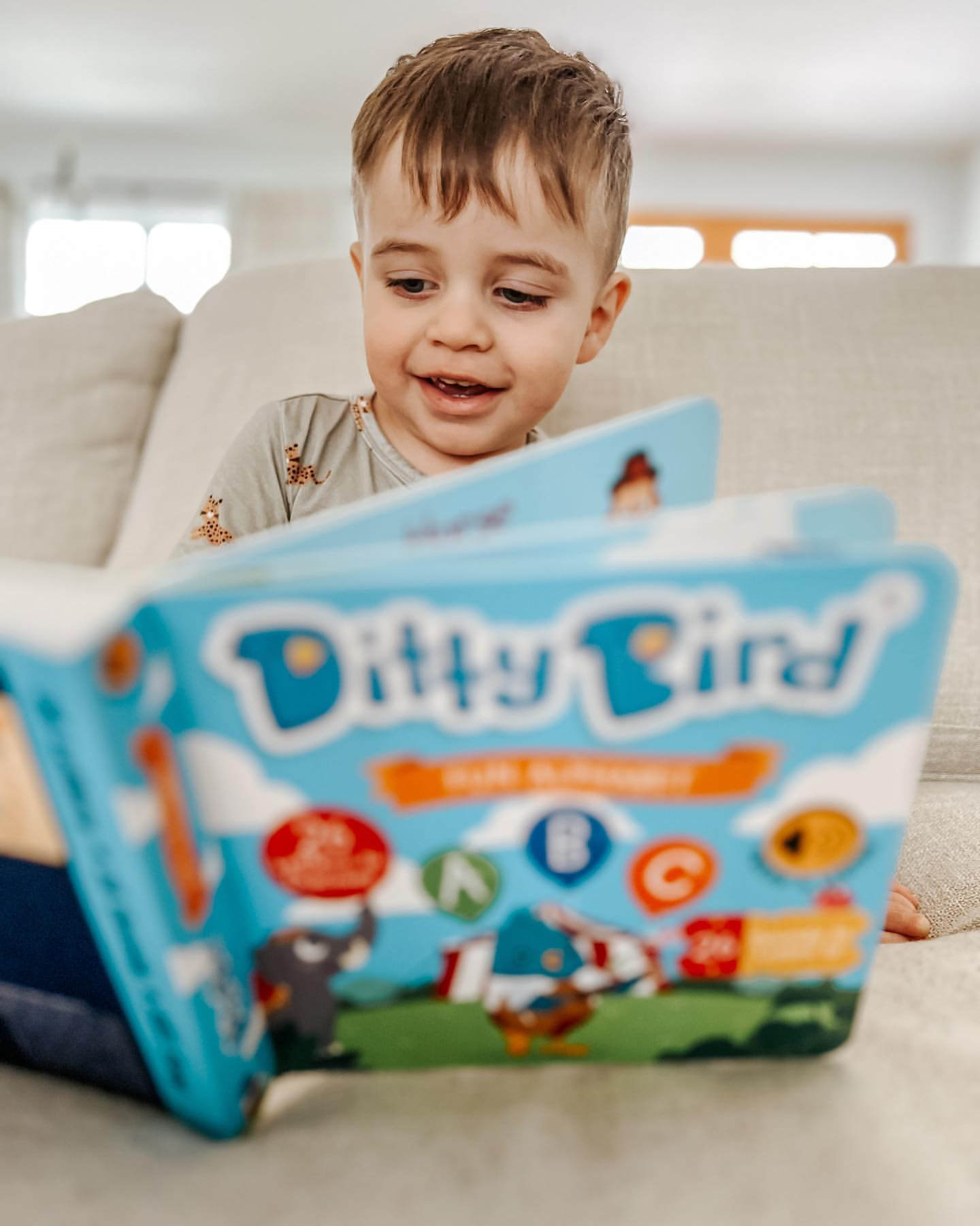A young child with light brown hair smiles on a sofa while exploring the open “ABC Fun Alphabet” interactive sound book by Ditty Bird UK, featuring colorful illustrations perfect for early learning and playful fun.