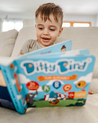 A young child with light brown hair smiles on a sofa while exploring the open “ABC Fun Alphabet” interactive sound book by Ditty Bird UK, featuring colorful illustrations perfect for early learning and playful fun.