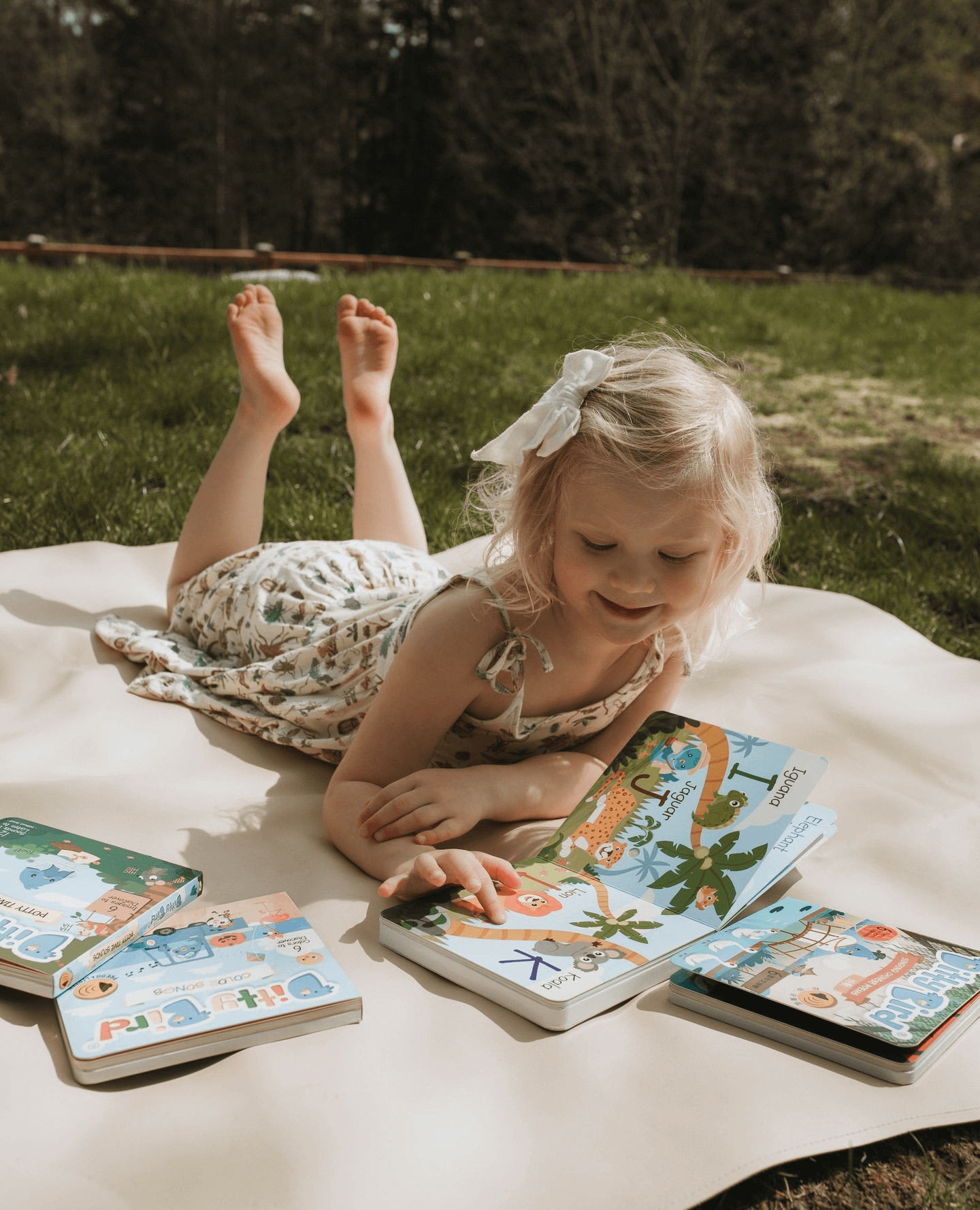 A young blonde girl with a white bow lies on a blanket outdoors, smiling as she looks at the Ditty Bird UK ABC Fun Alphabet book and early learning toys spread out on the grass in front of her.
