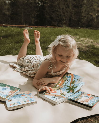 A young blonde girl with a white bow lies on a blanket outdoors, smiling as she looks at the Ditty Bird UK ABC Fun Alphabet book and early learning toys spread out on the grass in front of her.