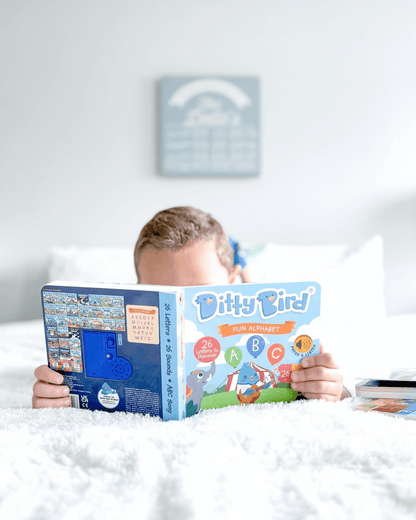 A child lies on a white bed, reading the Ditty Bird UK ABC Fun Alphabet book—an engaging interactive sound book and an ideal first birthday gift. The softly blurred background reveals a wall sign and more books to the right.
