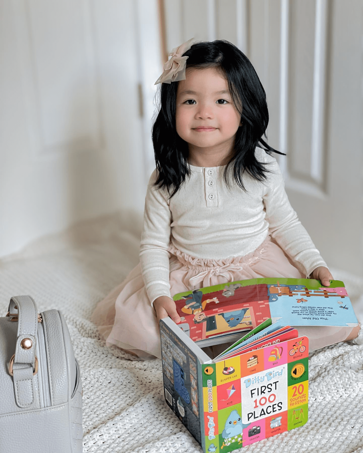 A young girl with long black hair and a pink bow sits on a white blanket, holding open Ditty Bird UK's Learning Songs book. A light gray handbag and an educational toy are next to her.