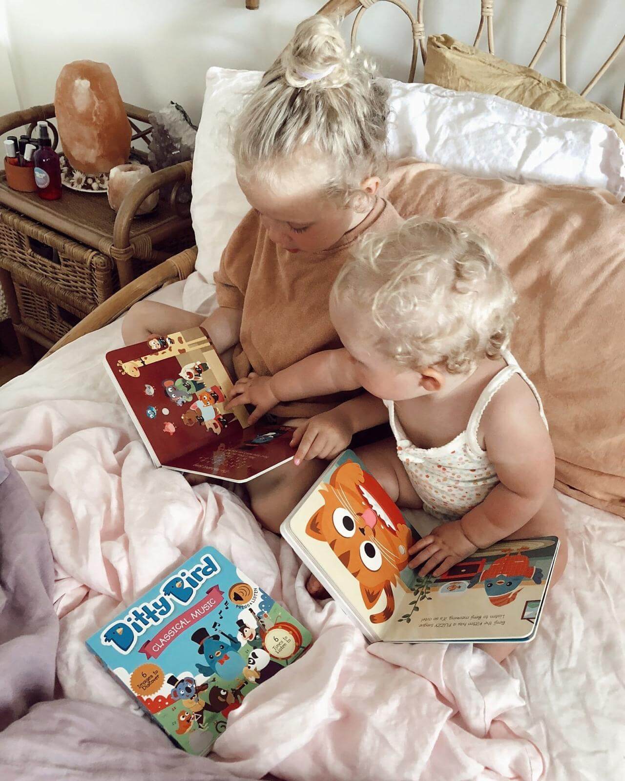 Two young blonde children sit on a bed reading colorful books. Nearby lies "Learning Songs" by Ditty Bird UK, one of the best musical toys for toddlers 1-3. The soft, neutral room offers a cozy space for early learning and play.