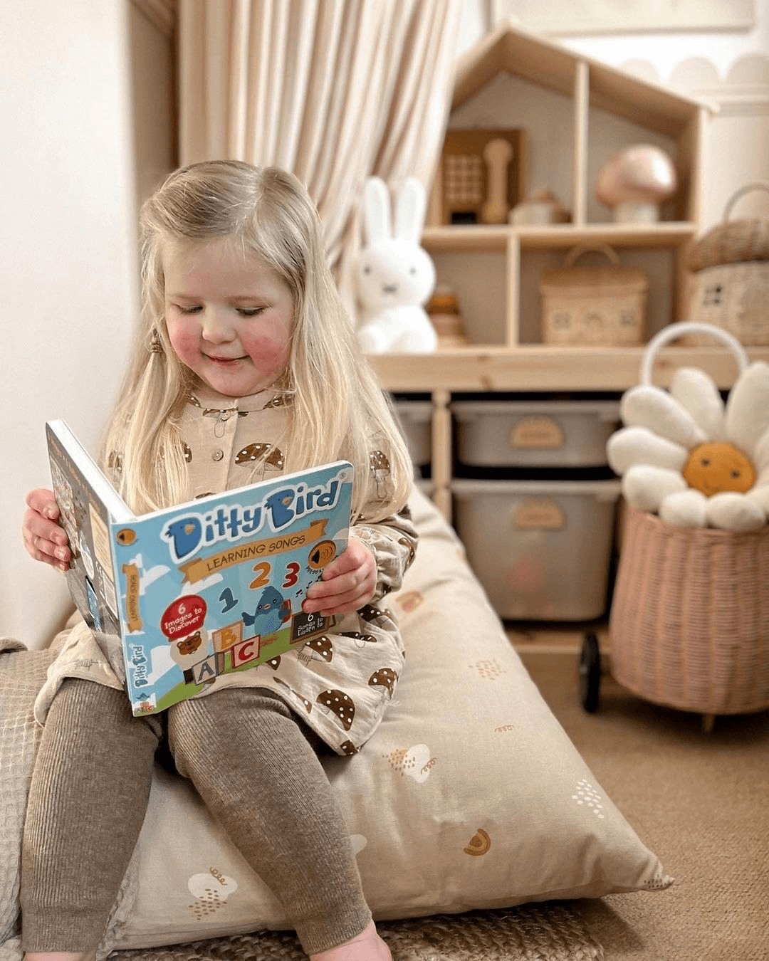 A young girl with long blonde hair smiles as she enjoys Ditty Bird UK's "Learning Songs" interactive sound book, surrounded by toys and a flower pillow—a charming scene that makes an ideal first birthday gift.