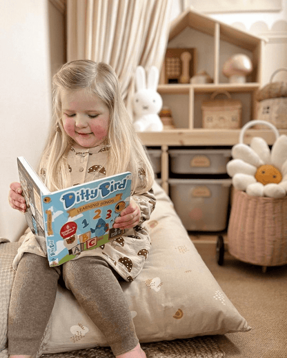 A young girl with long blonde hair smiles as she enjoys Ditty Bird UK's "Learning Songs" interactive sound book, surrounded by toys and a flower pillow—a charming scene that makes an ideal first birthday gift.