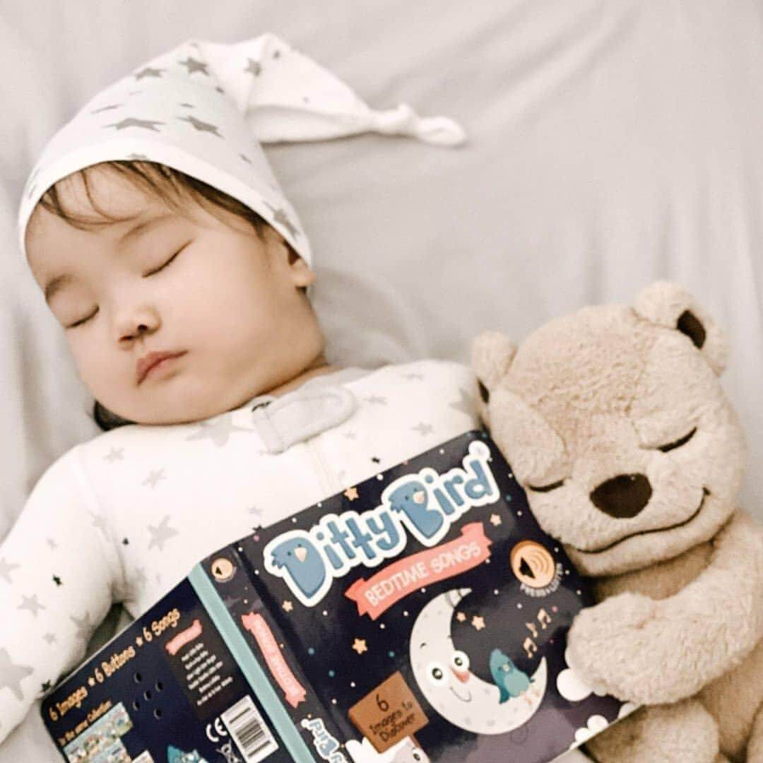 A baby in a star-patterned hat peacefully cuddles a beige teddy bear and holds Ditty Bird UK's Bedtime Songs interactive sound book while resting on a light blanket.