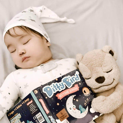 A baby in a star-patterned hat peacefully cuddles a beige teddy bear and holds Ditty Bird UK's Bedtime Songs interactive sound book while resting on a light blanket.