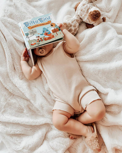 A baby lies on a cream blanket, holding Ditty Bird UK's Instrumental Songs sound book overhead. Surrounded by a pacifier and plush giraffe, this cozy early learning scene is perfect for a first birthday gift.