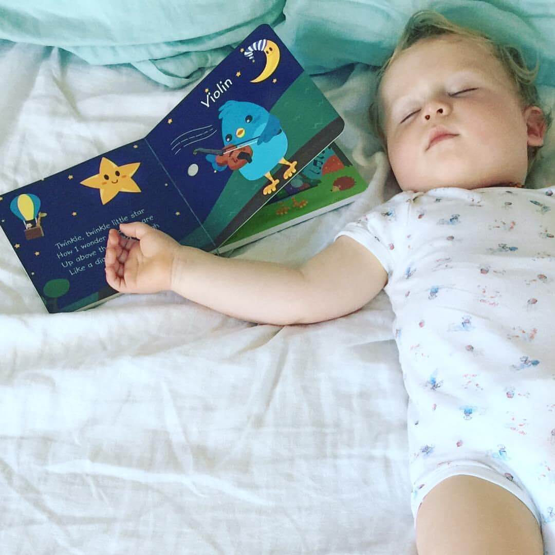 A toddler in a patterned onesie sleeps on a bed, holding Ditty Bird UK's "Instrumental Songs" interactive sound book—a delightful early learning toy with a blue bird playing violin and a smiling star, ideal for a first birthday gift.