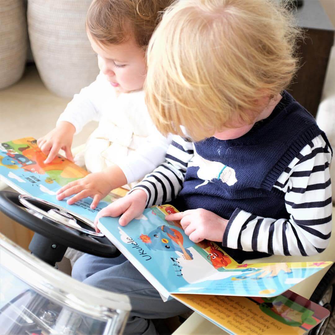 Two young children sit side by side in a toy car, exploring Ditty Bird UK’s Instrumental Songs book—an early musical learning moment. One wears a striped shirt and navy vest, the other a white top.