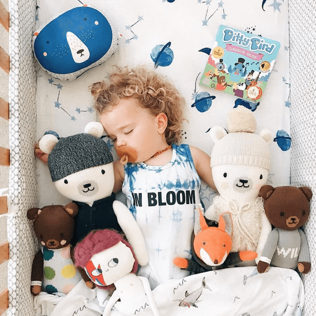 A toddler with curly blonde hair sleeps in a crib, surrounded by baby toys for 6–12 months, a plush blue bear pillow, and the Ditty Bird UK “Classical Music” book—perfect for early learning alongside cuddly friends. The child wears a tank top printed with IN BLOOM.