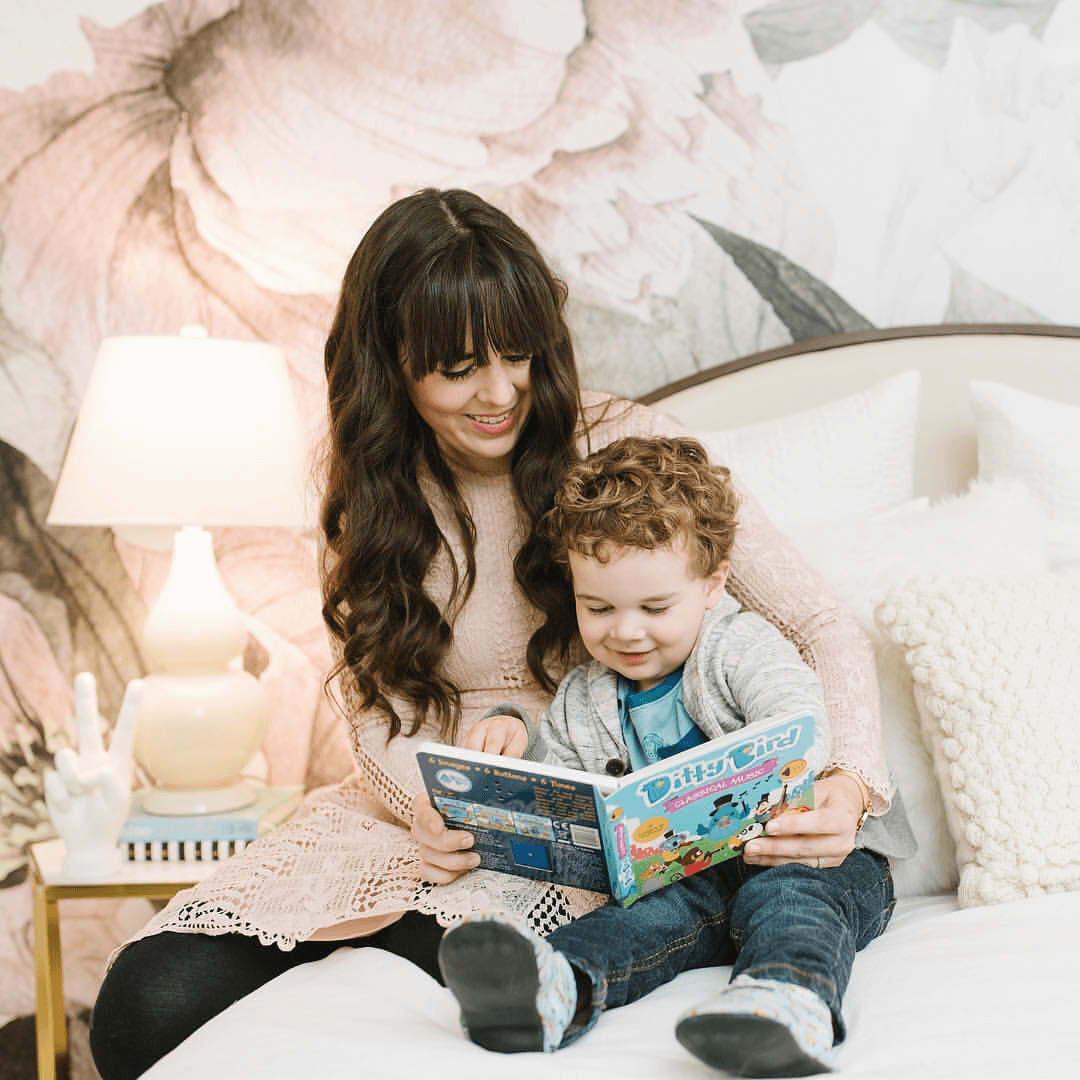 A woman and a smiling young boy sit on a bed reading the Ditty Bird UK "Classical Music" interactive sound book—an engaging musical toy for toddlers 1-3, perfect for sparking curiosity in a cozy setting.