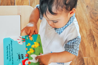 A young child in a white outfit with a blue-checkered collar stands on a wooden floor, pointing to ducks in the "Farm Animal Sounds" picture book by Ditty Bird UK—perfect for early learning fun.
