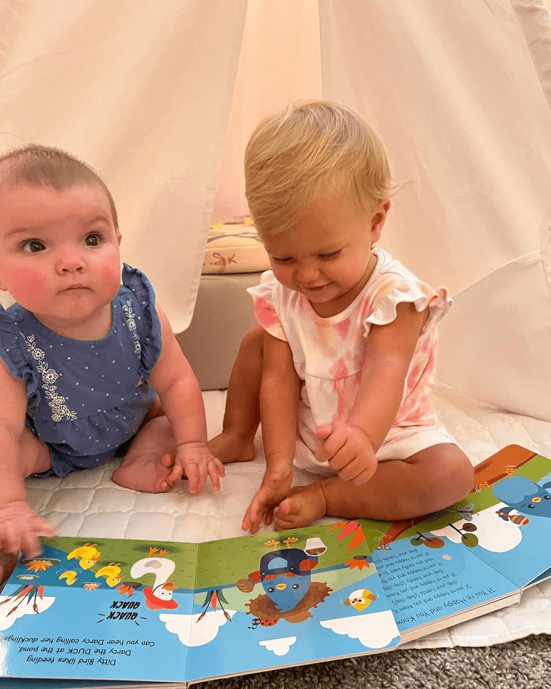 Two babies sit on a soft white mat inside a light-colored play tent. One, in blue, watches as the other, in pink-and-white, smiles and points at Ditty Bird UK's "Farm Animal Sounds" interactive sound book open before them.