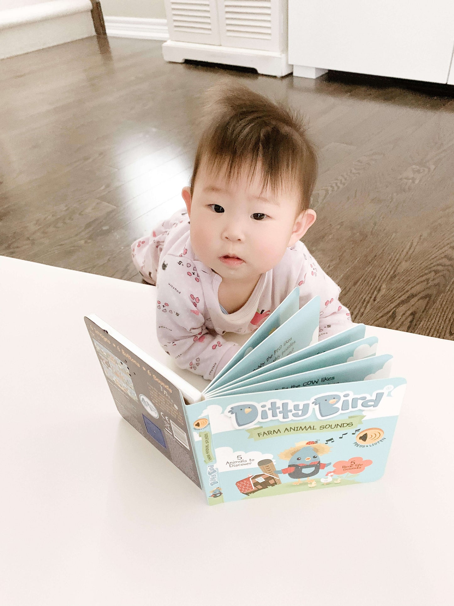 A baby in pajamas lies on a wooden floor, looking up while holding open the colorful Ditty Bird UK "Farm Animal Sounds" interactive sound book. White furniture is visible in the background.
