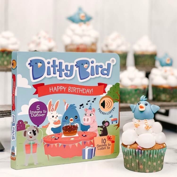 A Ditty Bird UK Happy Birthday book, the perfect first birthday gift and educational toy, stands next to a cupcake with a blue bird topper. Other white-frosted cupcakes appear blurred in the background on a light surface.