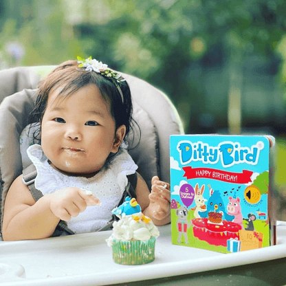 A smiling baby with cake on her face sits in a high chair, holding a cupcake with a blue bird topper. Beside her is Ditty Bird UK’s "Happy Birthday" book, the perfect fun and educational first birthday gift for little ones.