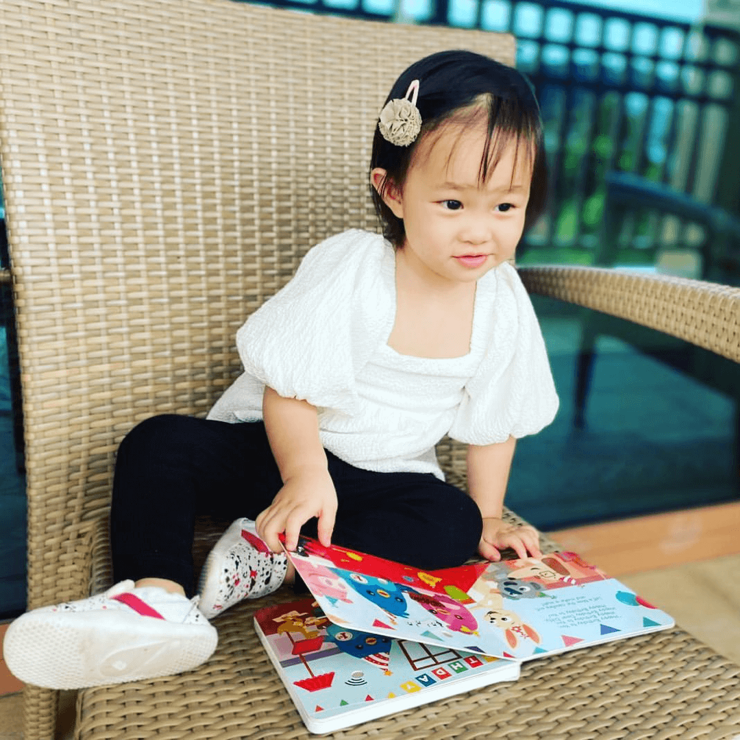 A young child with a hairclip sits on a wicker chair outdoors, smiling at the open Ditty Bird UK "Happy Birthday" interactive sound book on their lap—a delightful early learning pick or first birthday gift.