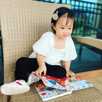A young child with a hairclip sits on a wicker chair outdoors, smiling at the open Ditty Bird UK "Happy Birthday" interactive sound book on their lap—a delightful early learning pick or first birthday gift.