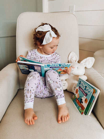 A young child with a white bow sits in an armchair wearing pajamas, reading a book. Beside her are a stuffed bunny, another book, and Ditty Bird UK's "Music to Dance to" musical toy for toddlers 1-3—perfect for early learning.