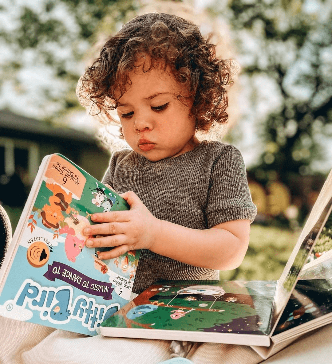 A young child with curly hair, in a gray shirt, sits outdoors and looks intently at Ditty Bird UK's Music to Dance to book—an educational toy ideal as a first birthday gift—with sunlight filtering through the trees.