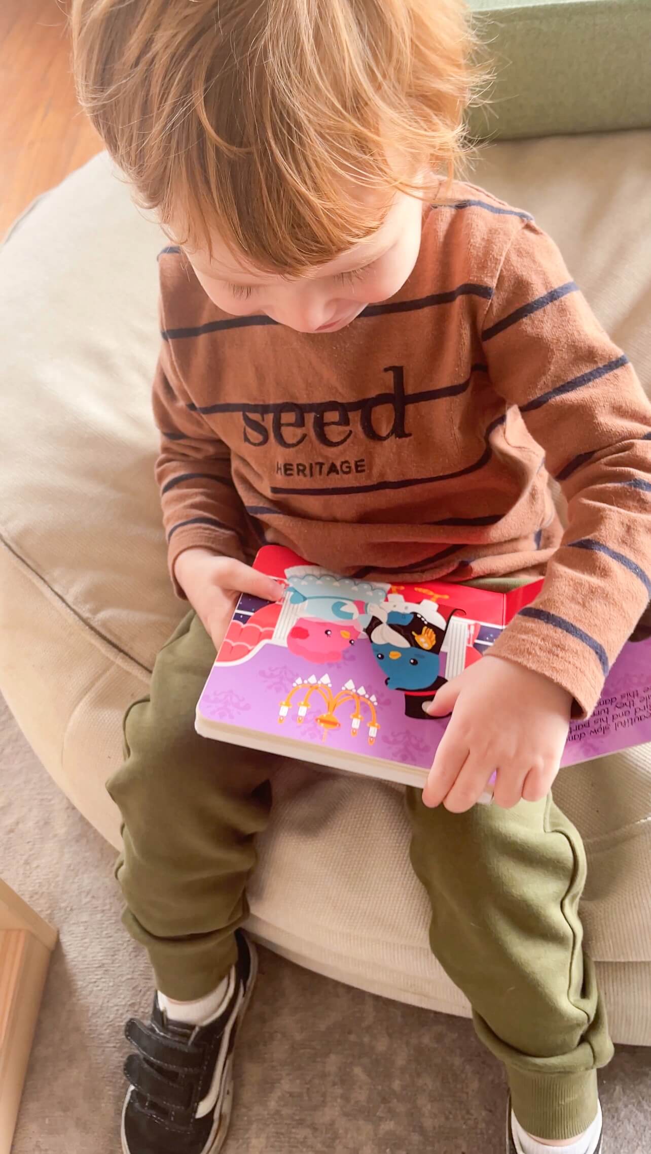 A young child with light brown hair sits on a beige chair, reading "Music to Dance to" by Ditty Bird UK. Dressed in a brown Seed Heritage shirt, green pants, and black sneakers—an ideal pick for early learning or a first birthday gift.
