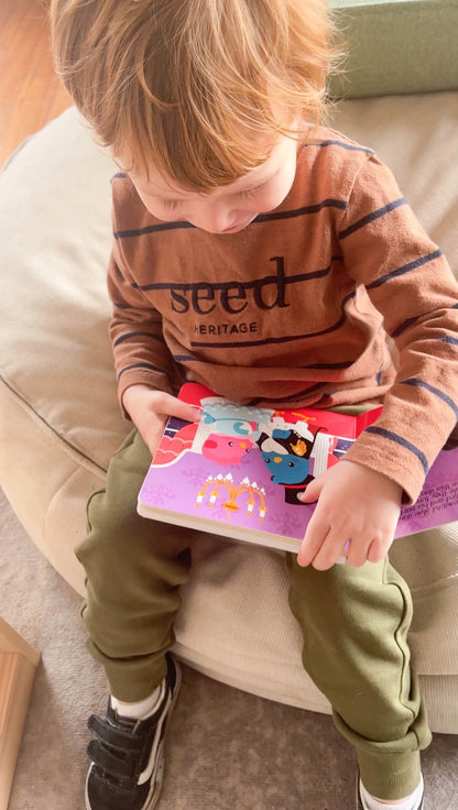 A young child with light brown hair sits on a beige chair, reading "Music to Dance to" by Ditty Bird UK. Dressed in a brown Seed Heritage shirt, green pants, and black sneakers—an ideal pick for early learning or a first birthday gift.