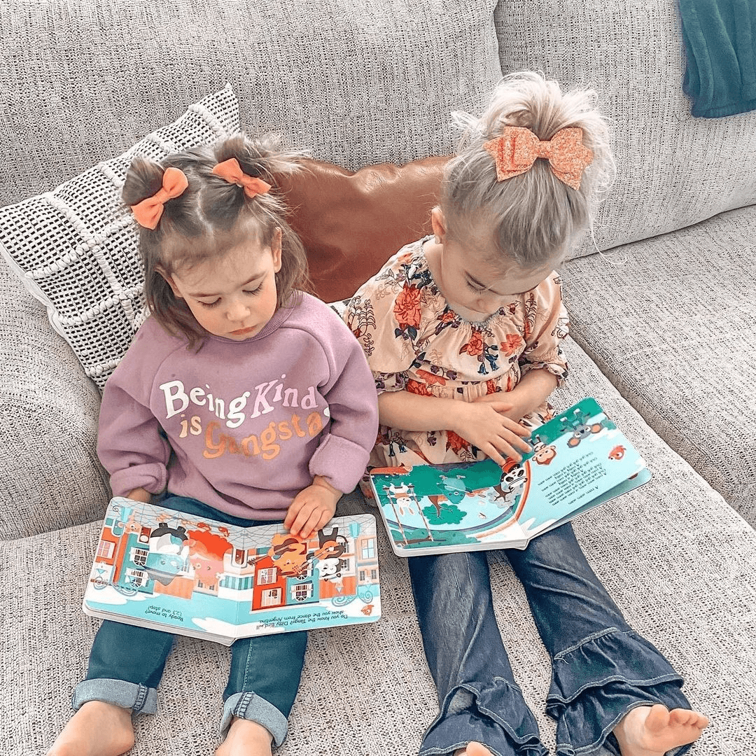 Two young girls sit on a light gray couch reading Ditty Bird UK's "Music to Dance to" book—perfect inspiration for choosing a first birthday gift or baby toys for ages 6-12 months.