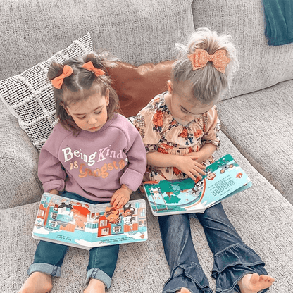 Two young girls sit on a light gray couch reading Ditty Bird UK's "Music to Dance to" book—perfect inspiration for choosing a first birthday gift or baby toys for ages 6-12 months.