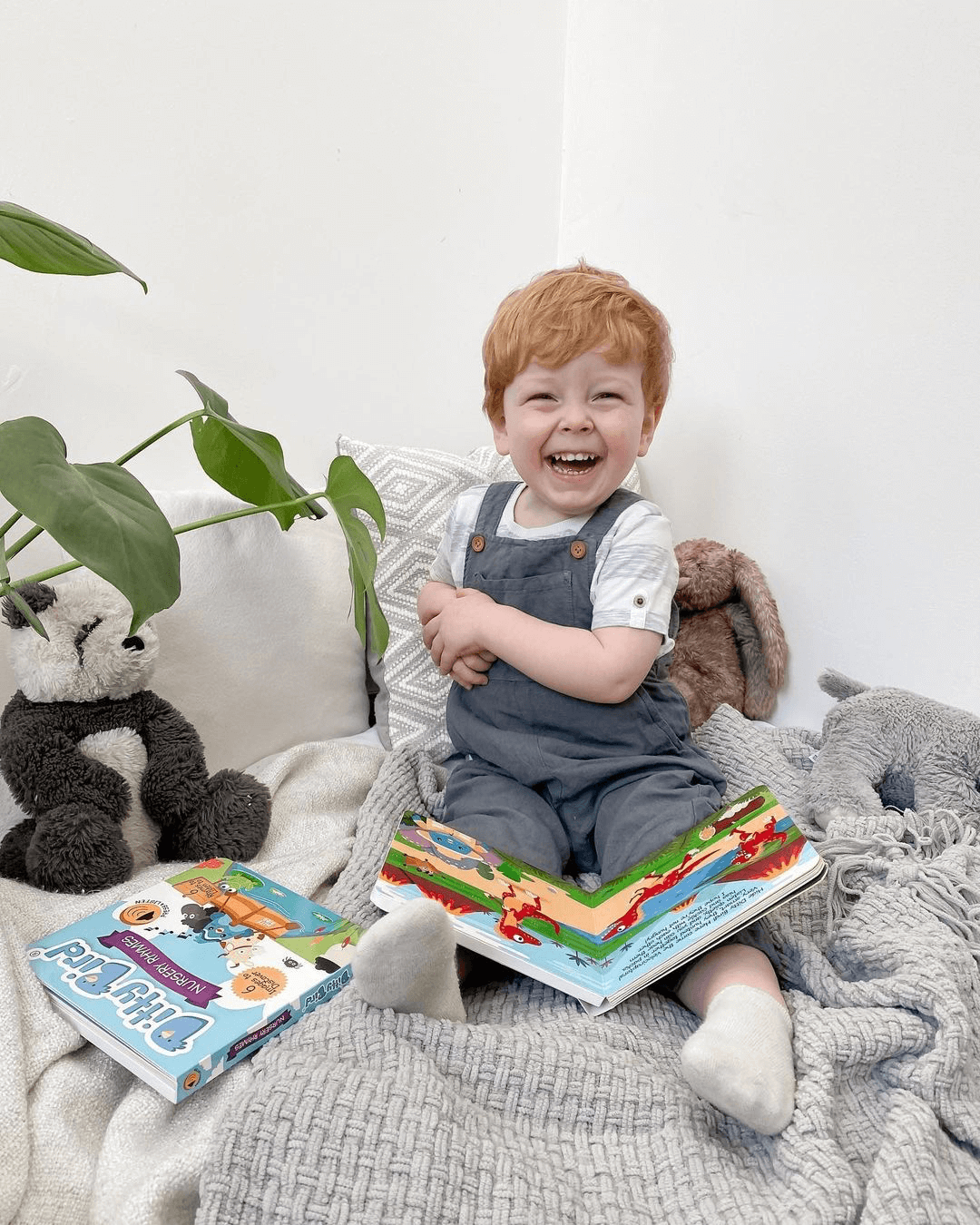 A smiling red-haired child sits on a cozy blanket holding Ditty Bird UK’s Dinosaur Sounds interactive sound book, surrounded by books, baby toys for 6-12 months, and stuffed animals. A green plant and sunlight brighten the cheerful room.
