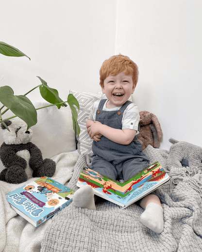 A smiling red-haired child sits on a cozy blanket holding Ditty Bird UK’s Dinosaur Sounds interactive sound book, surrounded by books, baby toys for 6-12 months, and stuffed animals. A green plant and sunlight brighten the cheerful room.