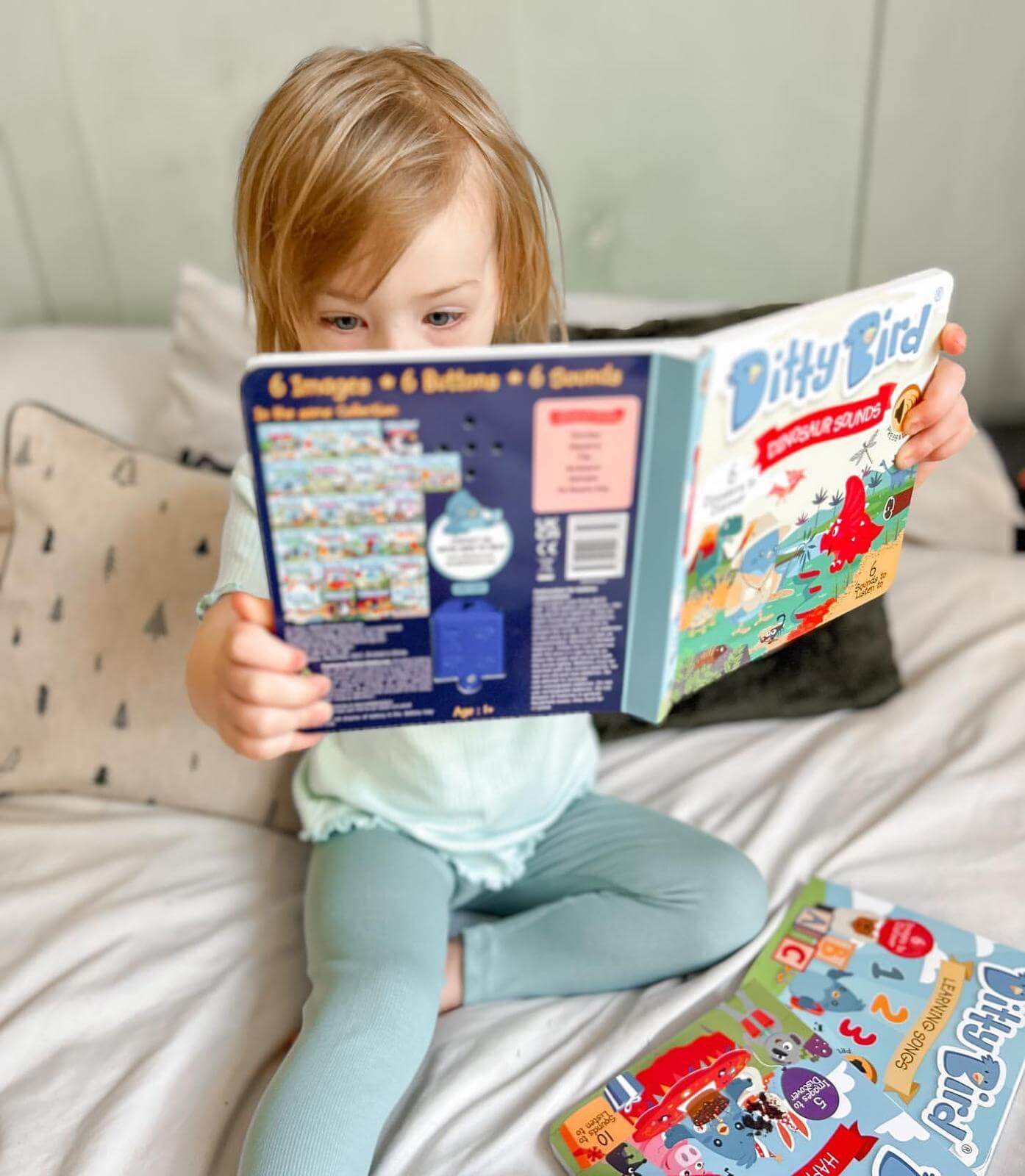 A young child with light hair sits on a bed, holding and reading Ditty Bird UK’s “Dinosaur Sounds” interactive sound book. Another book lies open nearby on the white bedding, with patterned pillows in the background.