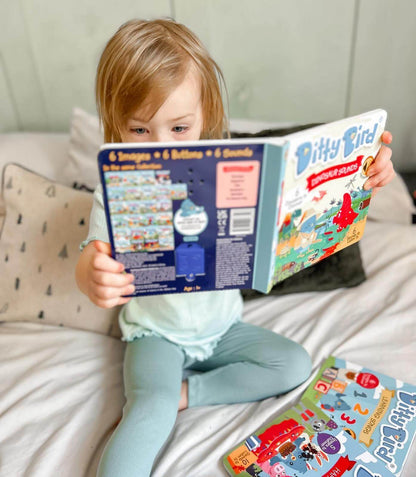 A young child with light hair sits on a bed, holding and reading Ditty Bird UK’s “Dinosaur Sounds” interactive sound book. Another book lies open nearby on the white bedding, with patterned pillows in the background.
