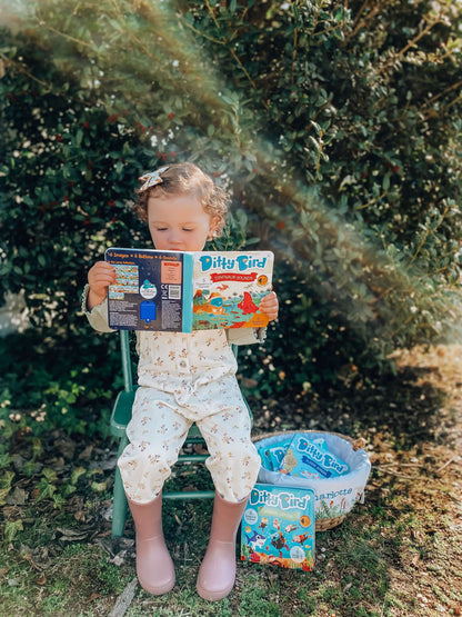 A young child sits on a green outdoor chair in floral pajamas and pink boots, reading Ditty Bird UK's "Dinosaur Sounds" interactive sound book. More Ditty Bird books sit in a basket nearby—great for early learning surrounded by nature.
